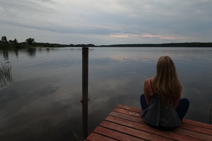 Woman with long blonde hair sits on a wooden dock overlooking a reflective lake under a cloudy, overcast dusk sky.