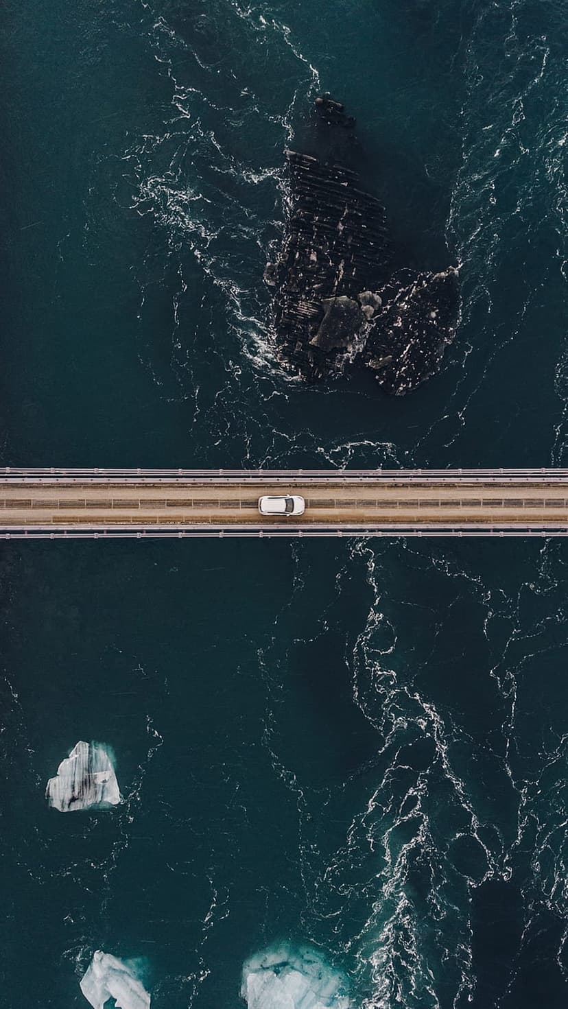 White car driving on a slender bridge over dark blue water with floating icebergs and a rocky shoreline cliff.