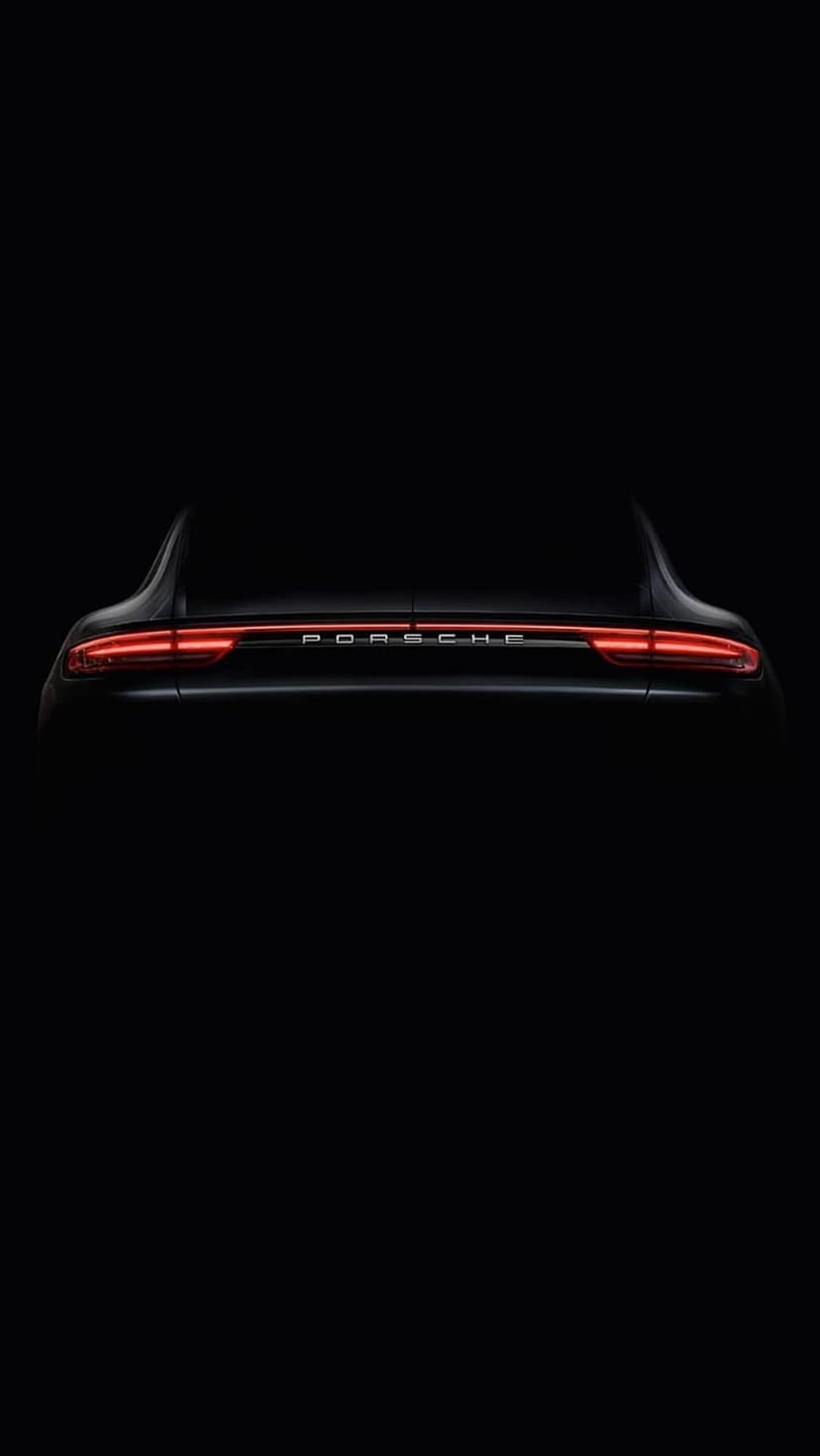 Red LED taillight strip on a black Porsche sports car glowing intensely within a dark, shadowed environment.