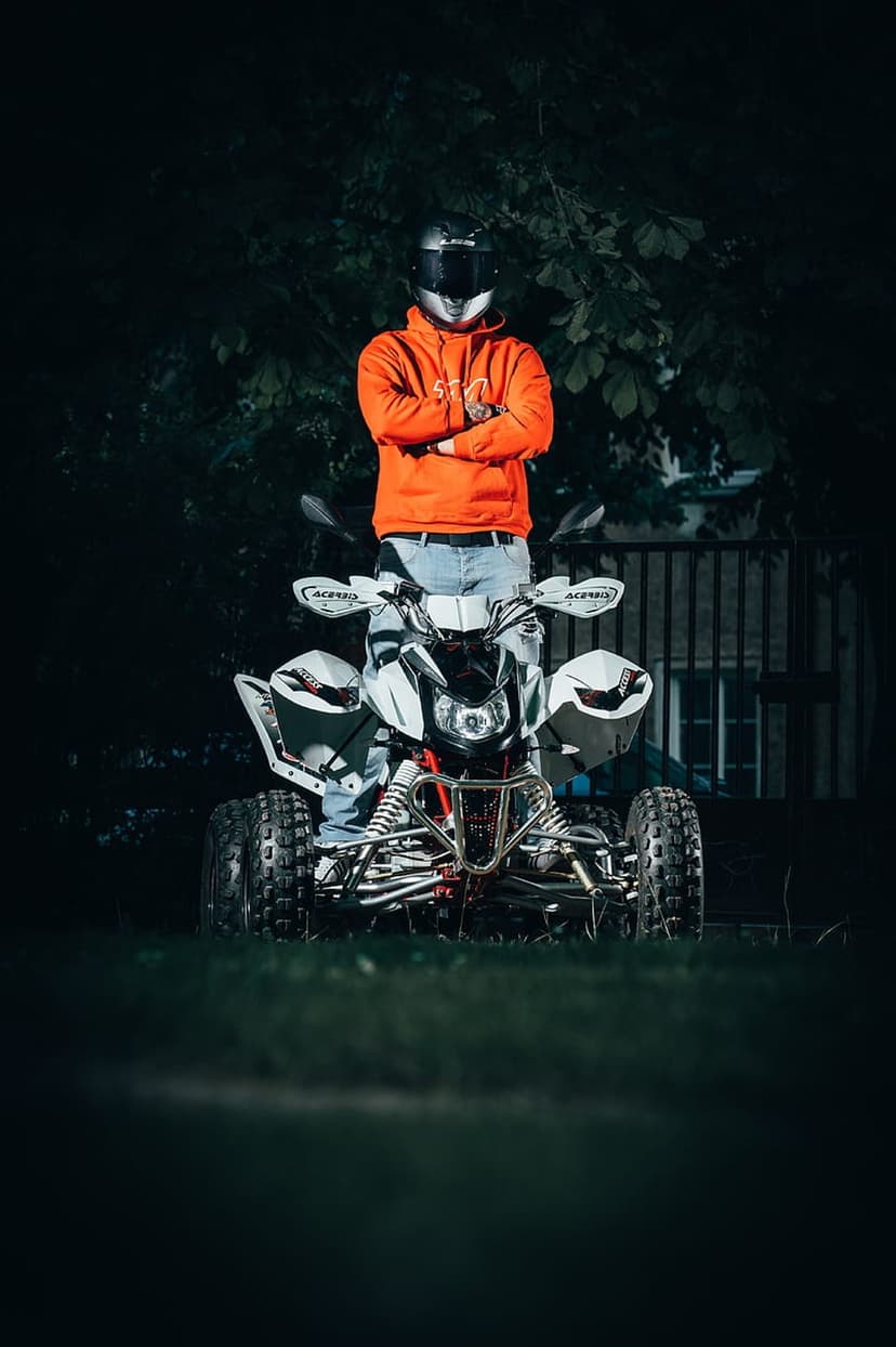 Person in an orange hoodie and helmet stands on a white ATV against a dark night background with a wooden fence.