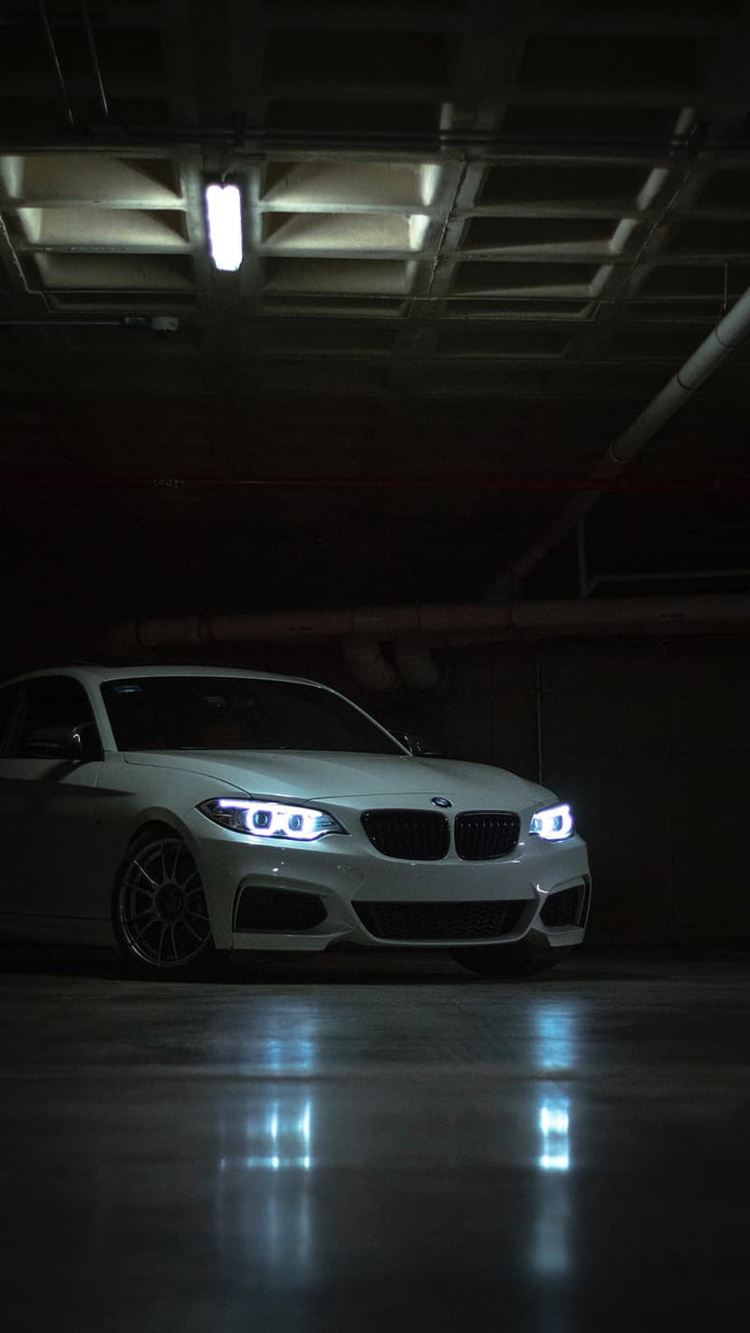 White BMW M2 with glowing LED headlights parked in a dark industrial garage with light reflecting on concrete.
