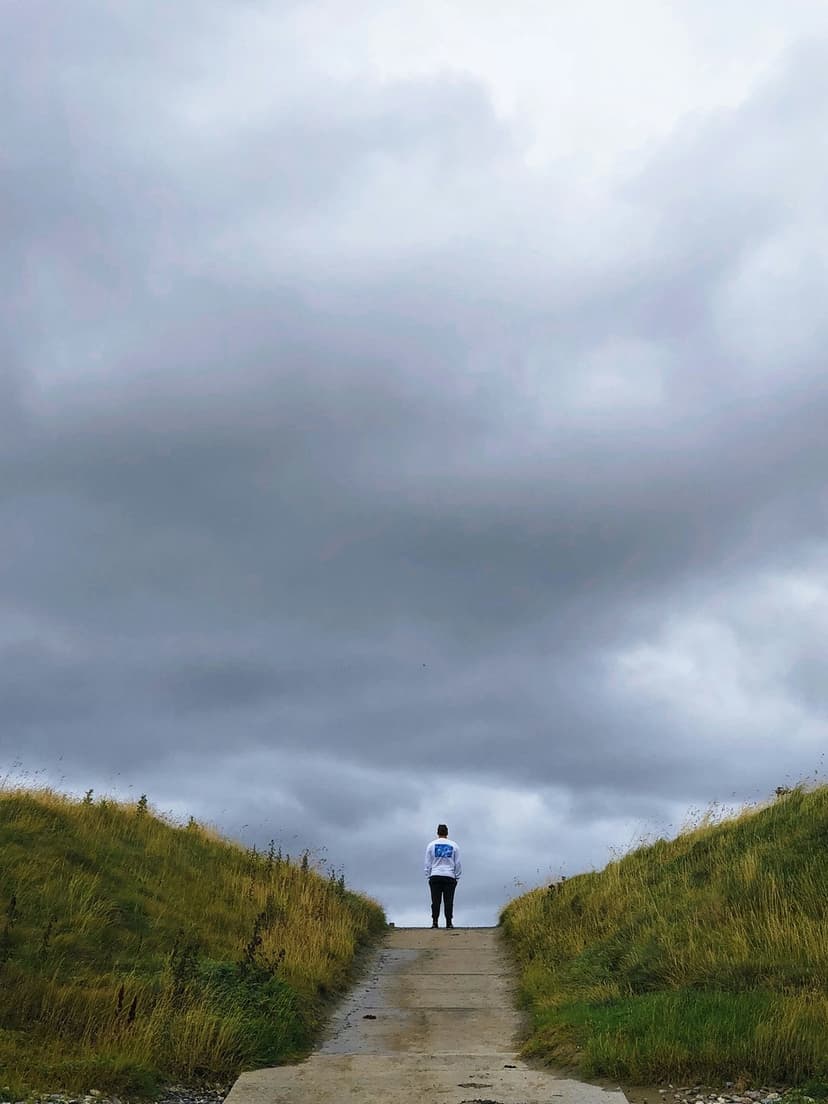 Lone figure on a concrete path faces a vast, dark stormy sky above vibrant green rolling hills in the distance.