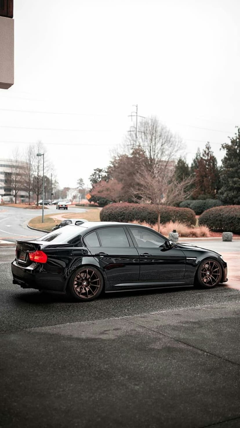 Black BMW sedan with bronze rims parked on asphalt under an overcast sky with trees and a building behind it.