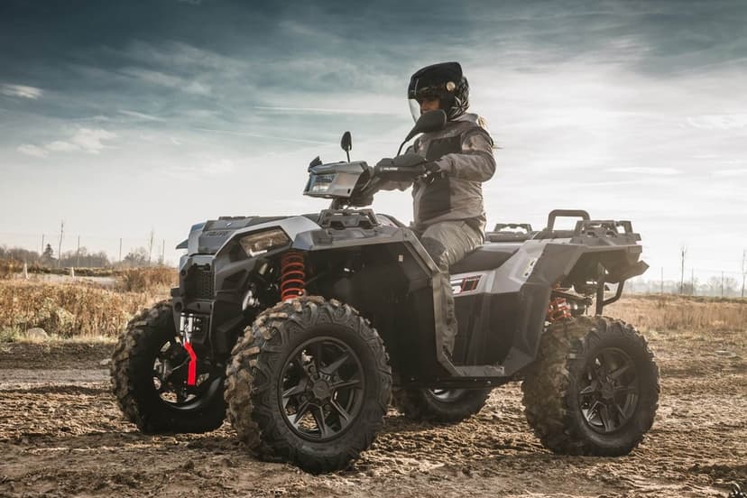 A rider on a grey Polaris ATV with orange springs sits in a muddy field under a hazy sky during the day.