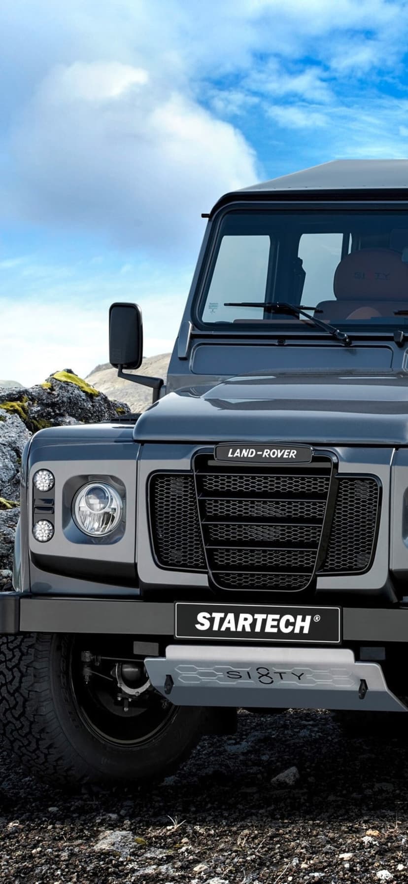 Close-up front view of a grey Land Rover Defender on rocky terrain under a bright blue sky with white clouds.