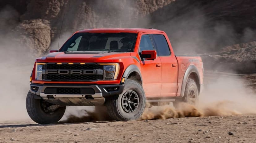 Orange Ford F-150 Raptor driving through a dusty desert landscape with rugged mountains in the background.