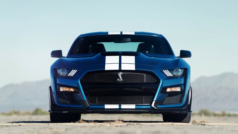 Blue Shelby GT500 muscle car with white racing stripes and glowing headlights against a desert background.