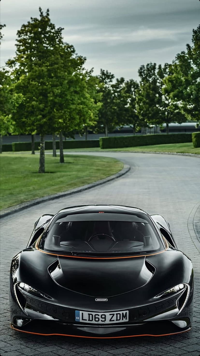 Black McLaren P1 supercar with orange accents on a stone driveway surrounded by green hedges and trees.