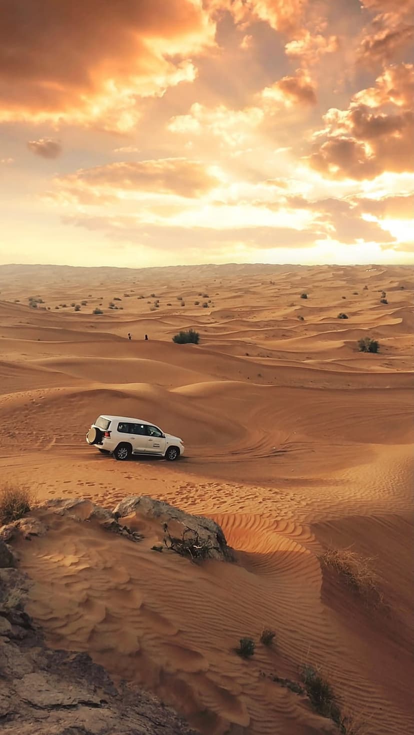 White SUV climbing a steep sand dune under a golden sunset sky with scattered clouds and desert shrubs.