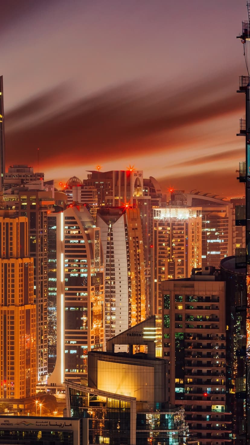 Towering Dubai skyscrapers glow with window lights against a reddish-orange twilight sky in a vertical view.