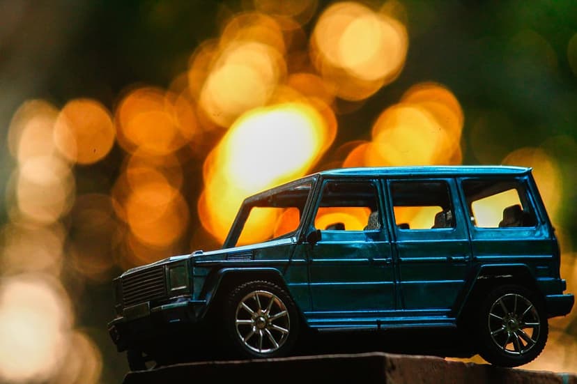 Teal toy SUV with sharp grille details sits before a blurred background of orange sunset bokeh lights.