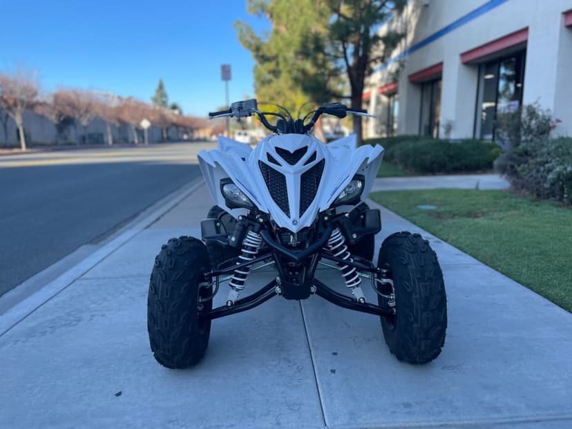 White Yamaha Raptor sport ATV parked on a sidewalk under a clear sky with visible suspension and large tires.