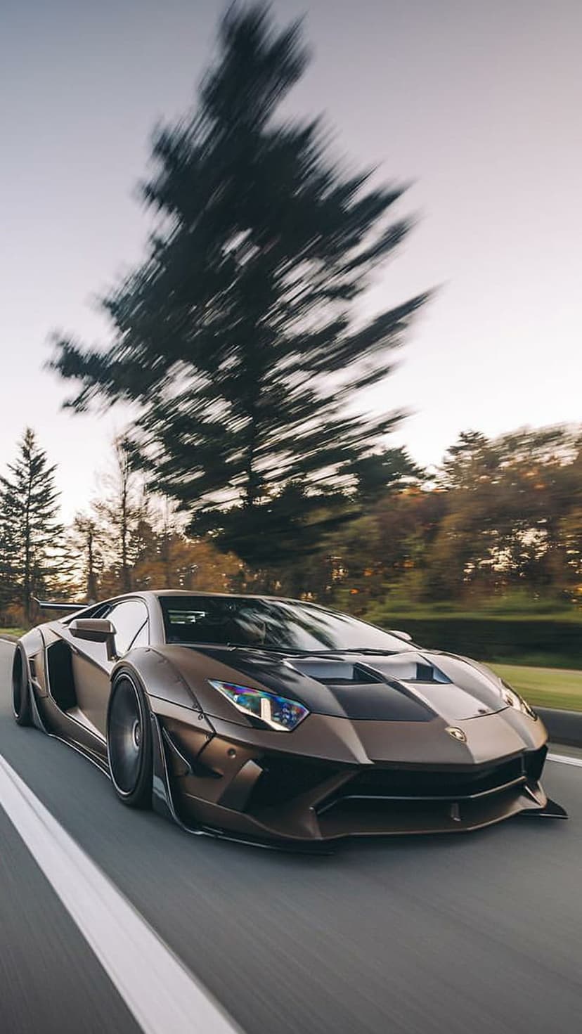 Brown Lamborghini Aventador with widebody modifications driving fast on a road with motion blur and trees.