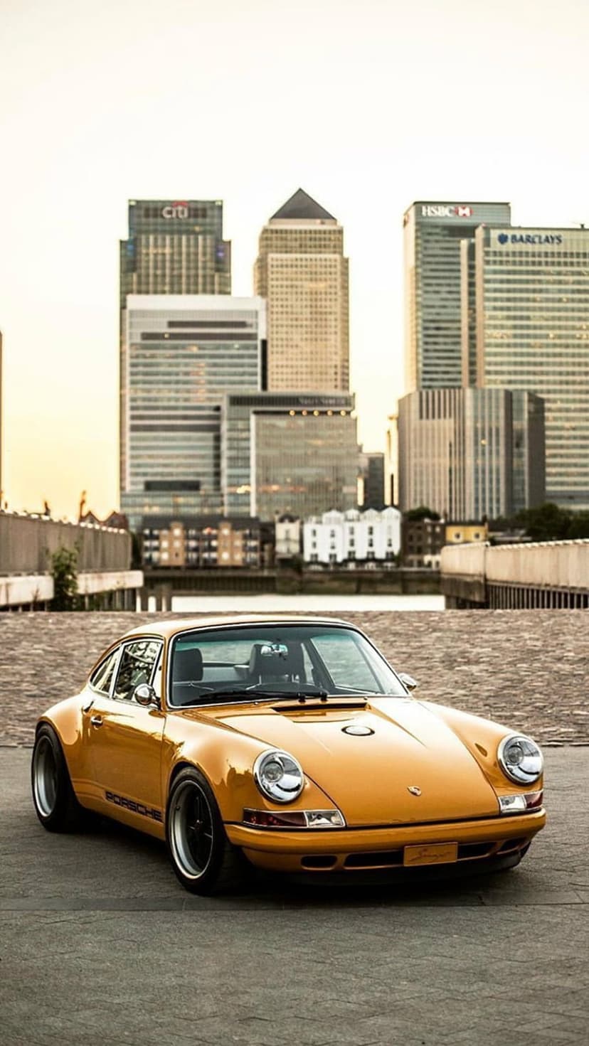 Orange vintage Porsche 911 parked on a paved street with the modern glass towers of Canary Wharf in the background.