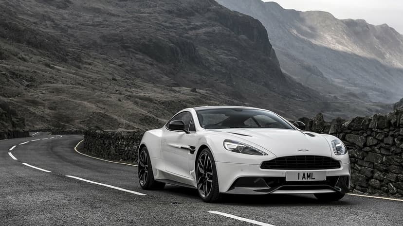 White luxury sports car parked on a winding mountain road with rugged rocky peaks and a rustic stone wall.