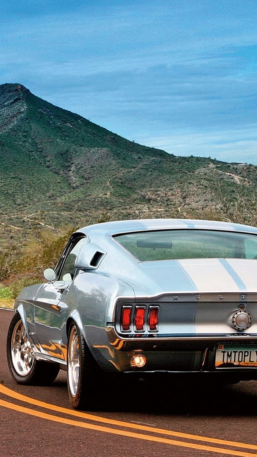Blue vintage Ford Mustang with white stripes drives on a desert road toward mountains under a clear sky.