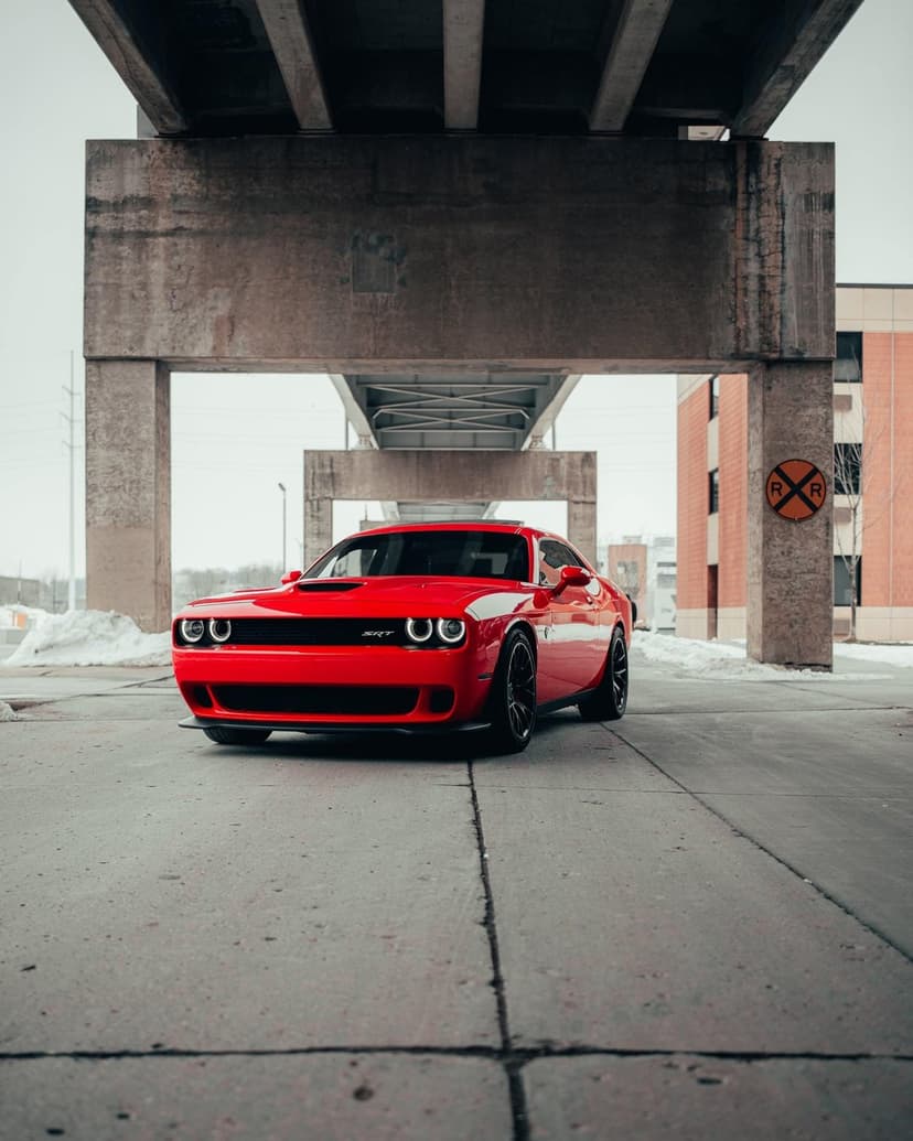 Red Dodge Challenger SRT muscle car parked under a gray concrete overpass in a winter urban street setting.