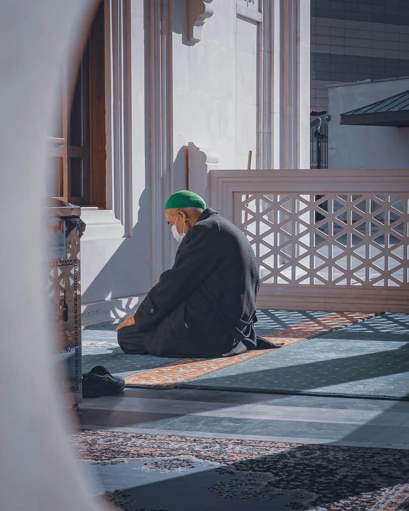 Man in a green hat kneels on a prayer rug inside a mosque with sunlight casting geometric shadows on the floor.