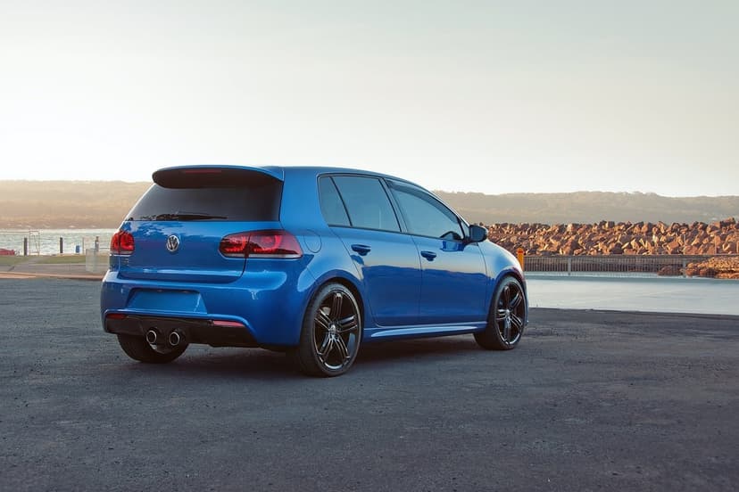 Blue Volkswagen Golf R parked on grey asphalt near a rocky coast and calm water under a soft golden hour sky.