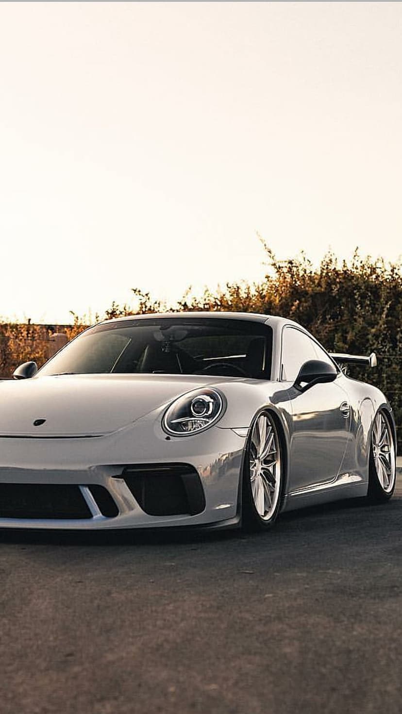 Silver Porsche 911 GT3 parked on asphalt with a rear wing, green trees, and a warm sky in the background.