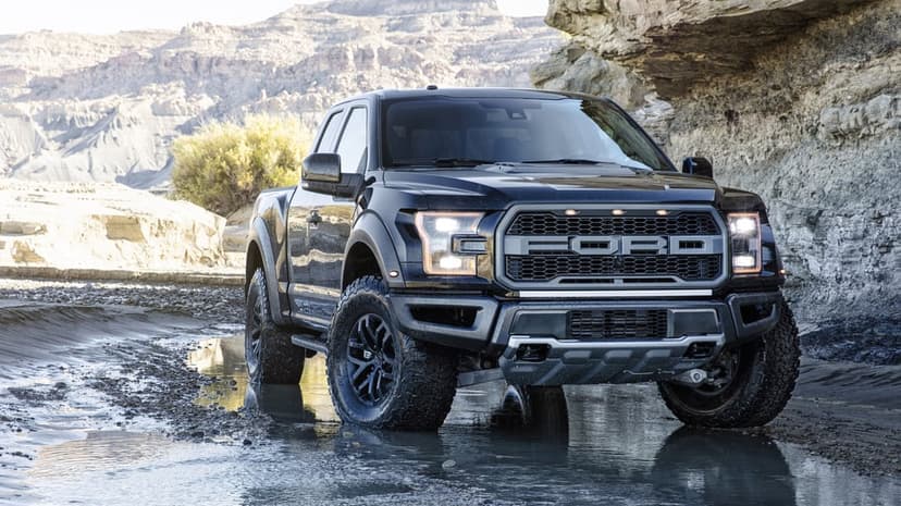 Black Ford F-150 Raptor truck parked in a shallow stream between rocky canyon walls under bright daylight.