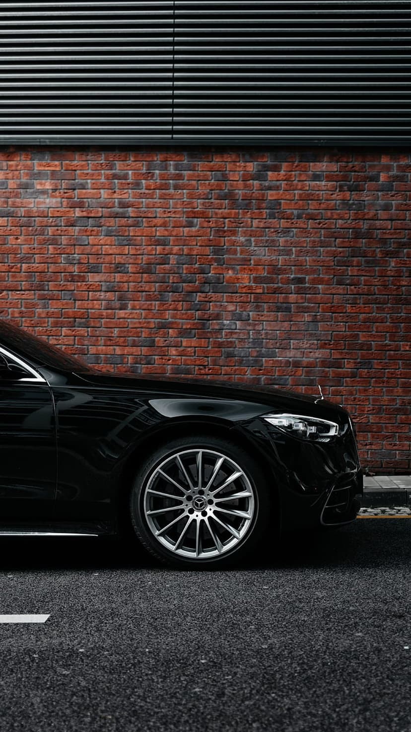 Black Mercedes-Benz S-Class front wheel and bumper parked on asphalt by a red brick wall and metal siding.