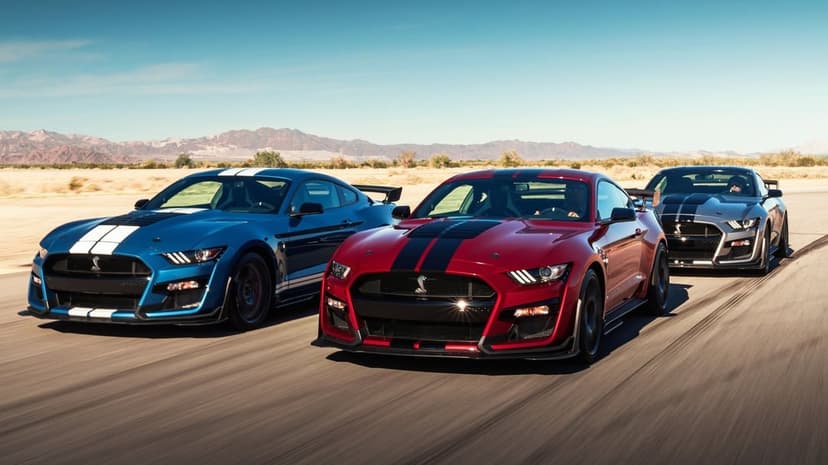 Blue, red, and grey Ford Mustang Shelby GT500 cars racing on a desert track with mountains under a blue sky.