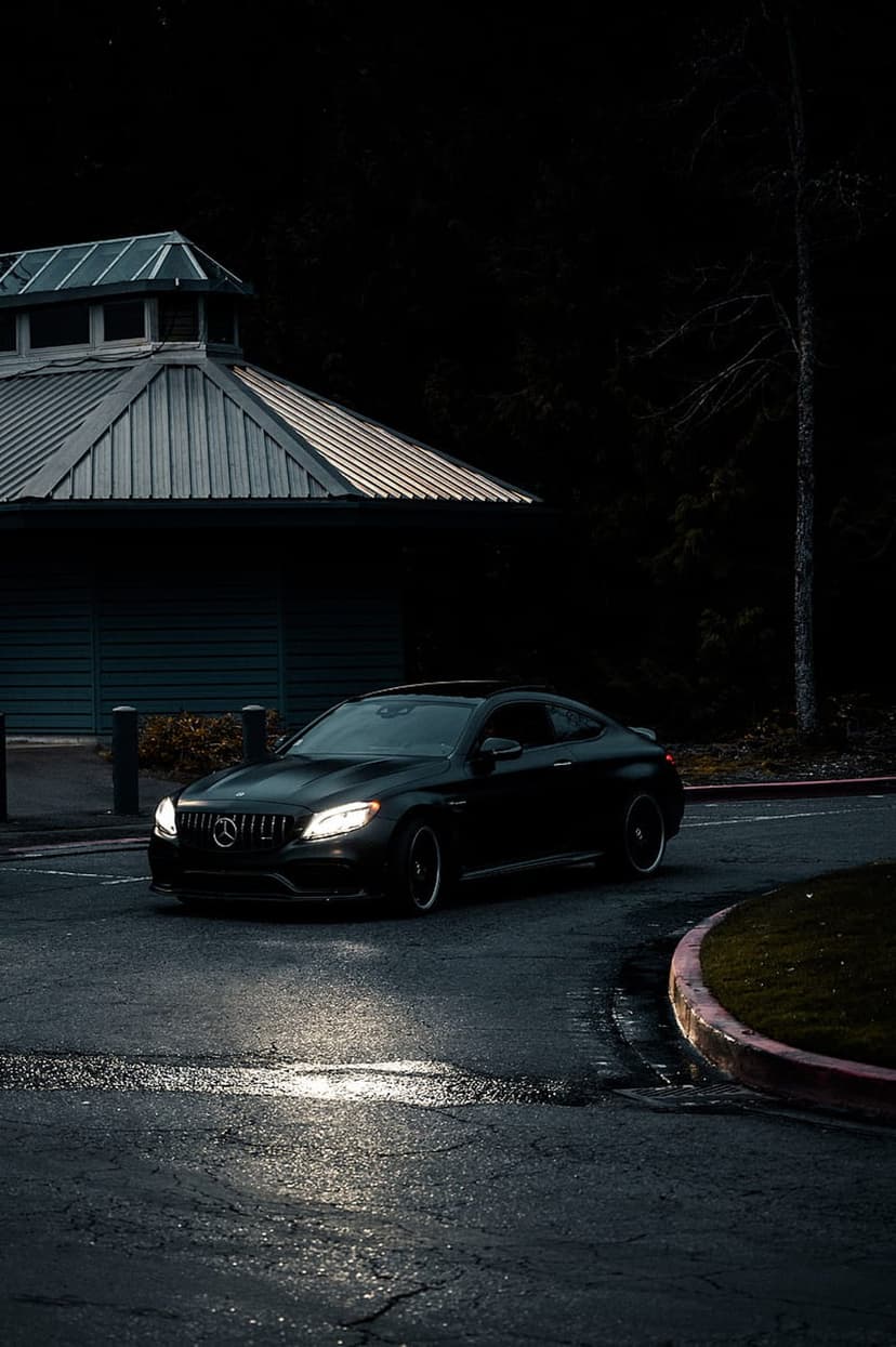Black Mercedes coupe sits on a wet road at twilight with headlights reflecting in puddles near a modern building.