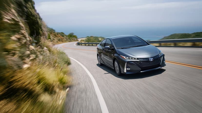 Dark gray Toyota Prius Prime driving on a winding coastal road with motion blur and ocean background.