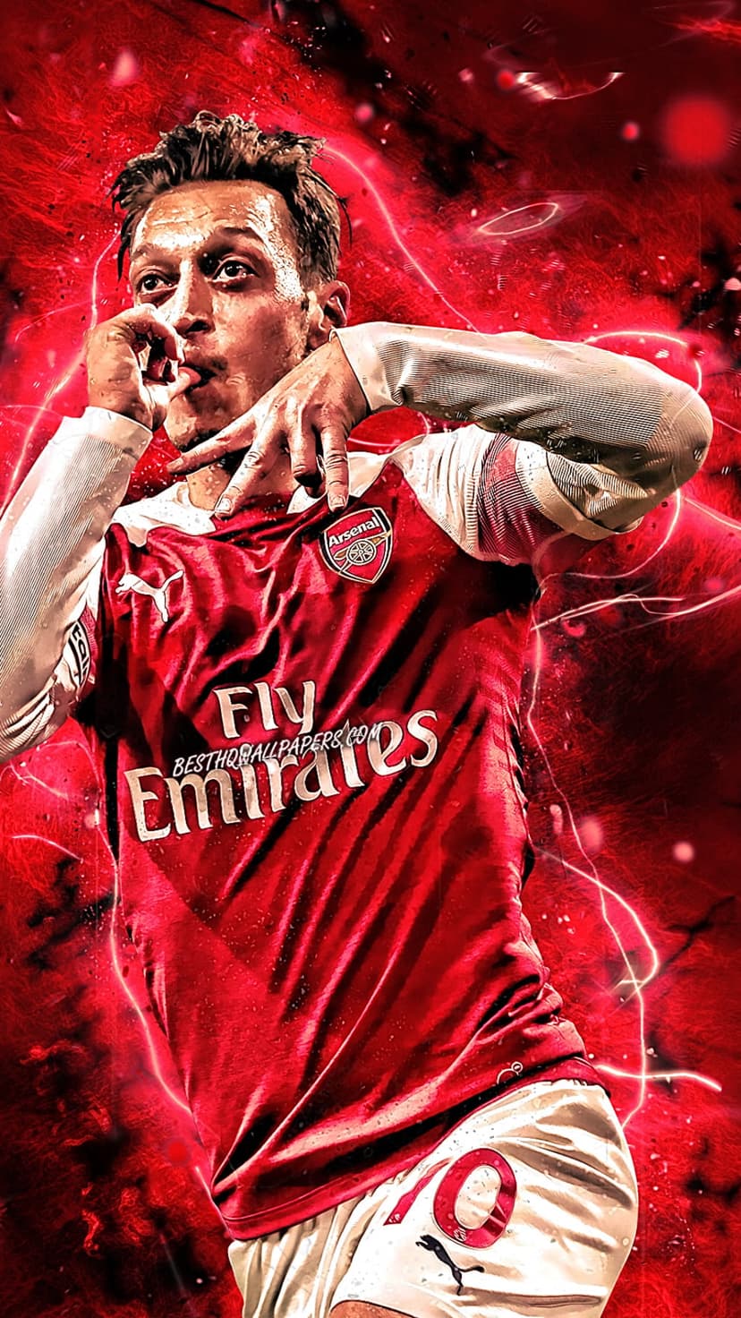 Mesut Ozil wearing a red Arsenal jersey stands on a grass football pitch with blurred stadium lights behind him.