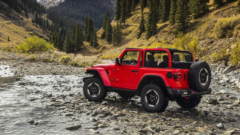 Red Jeep Wrangler Rubicon parked in a shallow rocky riverbed with steep pine-covered mountains in the background.