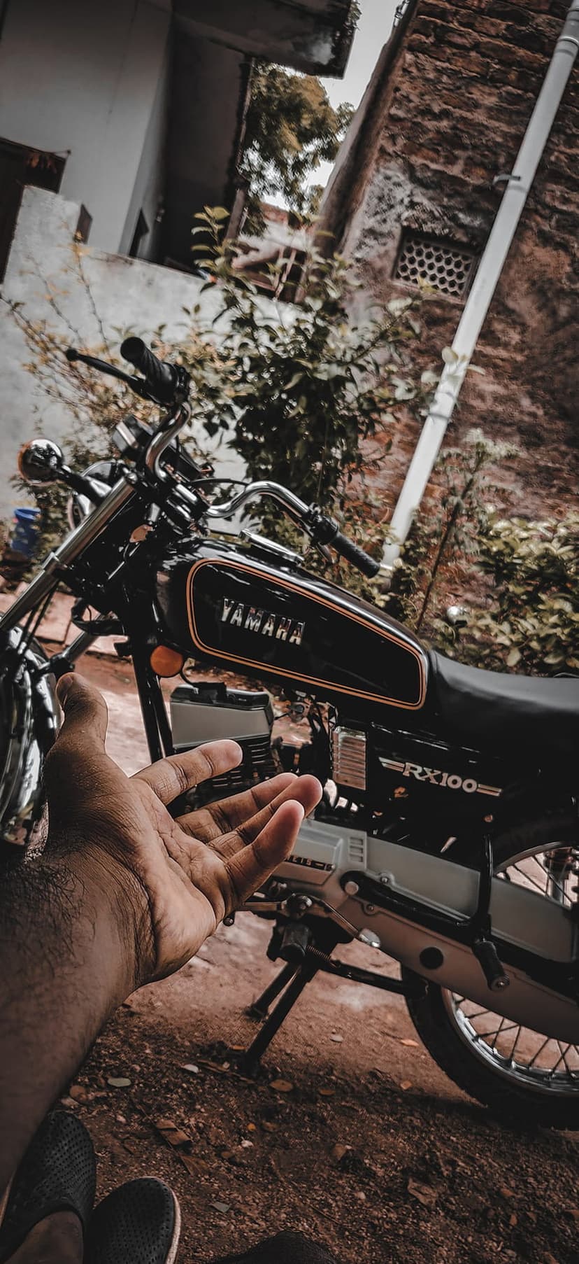 Black Yamaha RX100 motorcycle parked near a rustic building with a person's hand reaching toward the fuel tank.