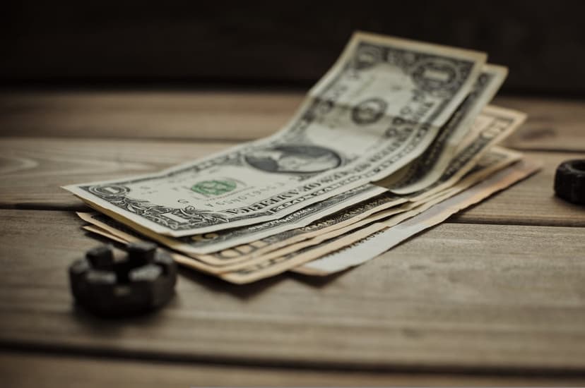 Fanned US dollar bills and a small metallic nut sit on a textured rustic wooden table under soft warm lighting.