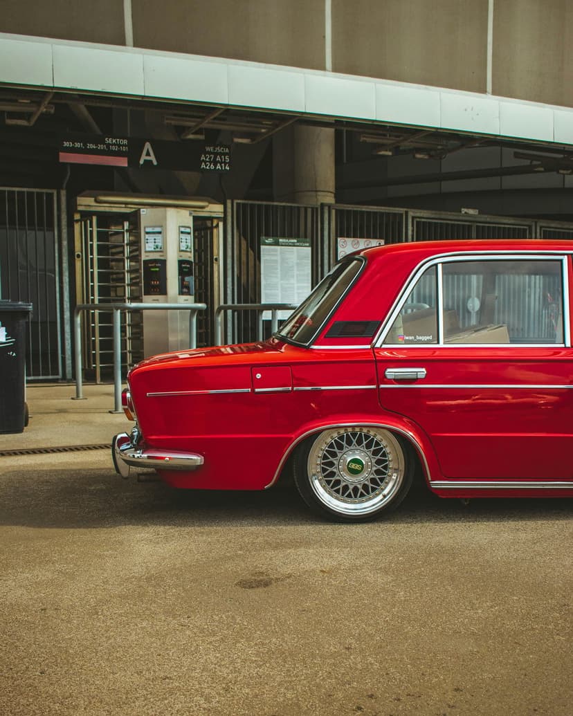 Red vintage sedan with lowered suspension and BBS wheels parked in front of a modern urban building entrance.