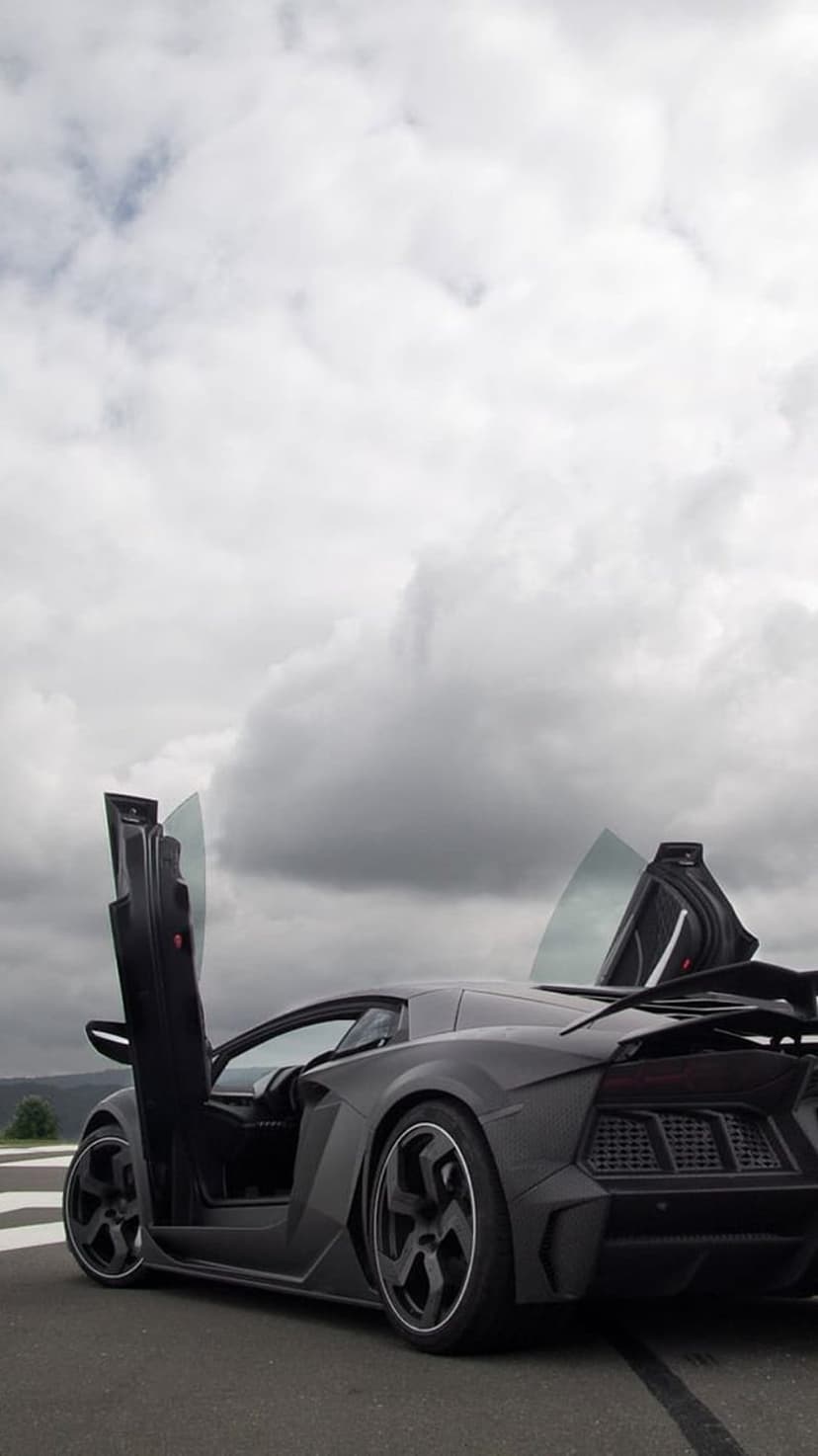Matte black Lamborghini Aventador with open scissor doors on a race track under a cloudy, overcast grey sky.