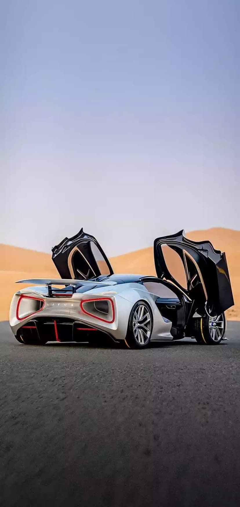 Silver supercar with open butterfly doors and red rear accents parked on a desert road near sand dunes.