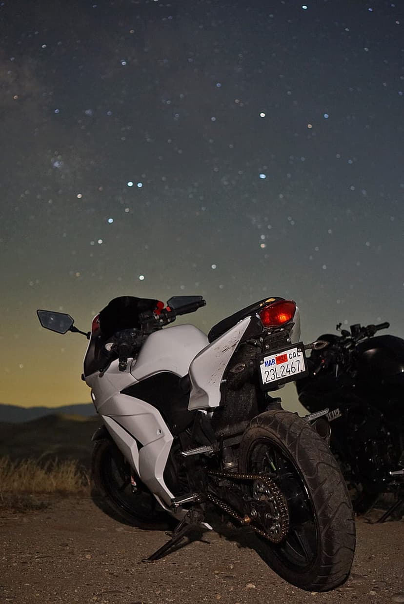 White sport motorcycle parked on a dirt path under a vivid Milky Way galaxy with stars and distant silhouettes.