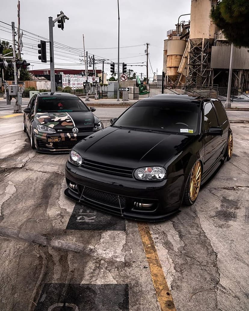 Two modified Volkswagen Golf MK4 hatchbacks parked at a railway crossing with industrial buildings in background.