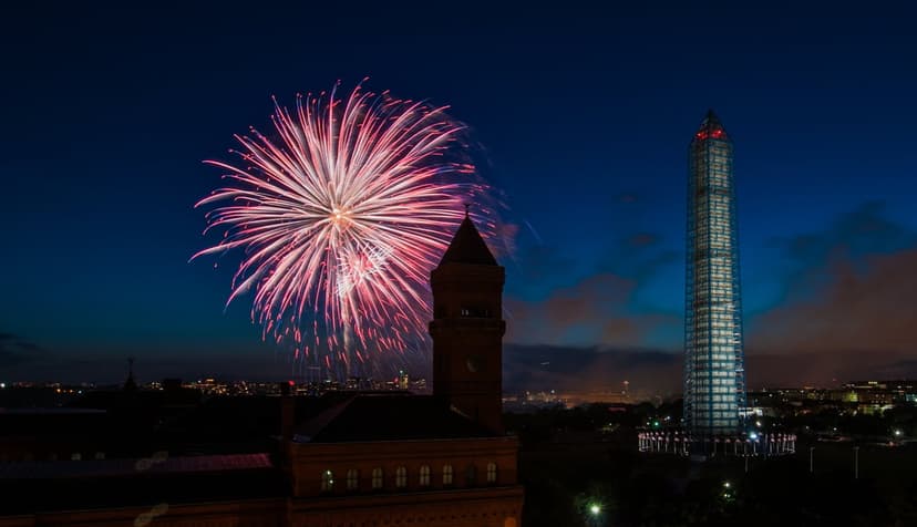 Washington Monument stands under pink and white fireworks with a city skyline and clock tower in the dark.