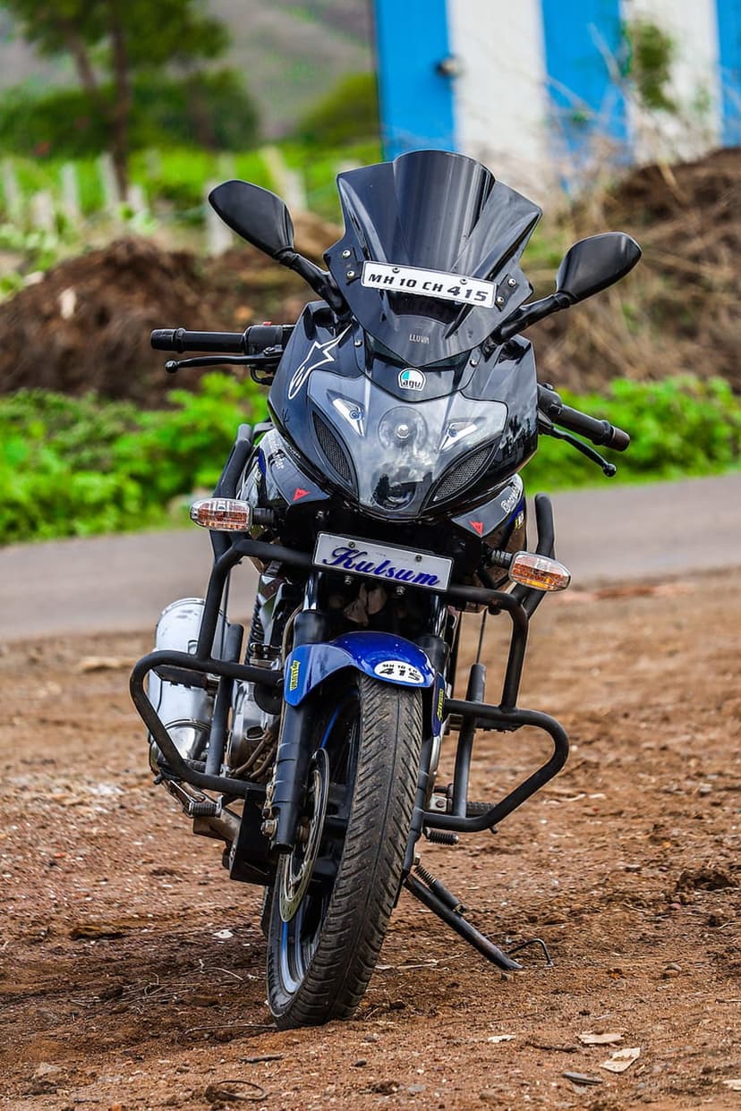 Blue Bajaj Pulsar motorcycle with chrome accents parked on a dusty rural road against blurred green foliage.