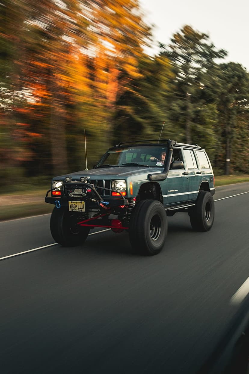 Dark green Jeep Cherokee XJ with off-road tires driving past blurred orange autumn trees on a paved road.