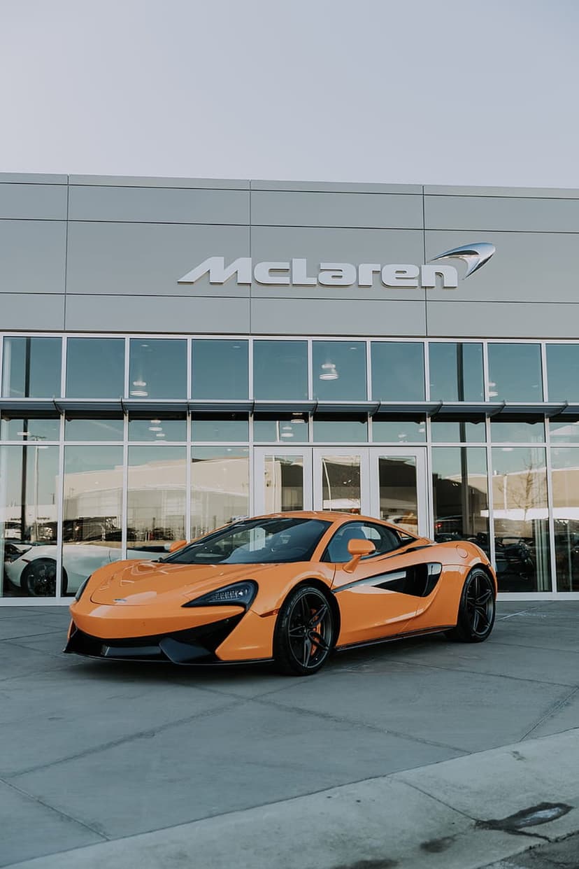 Orange McLaren 570S sports car with black wheels parked outside a modern glass dealership under a clear sky.