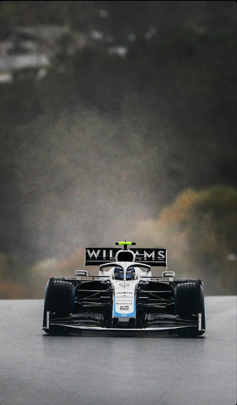Front wing of a blue and white Williams F1 car rests on a damp race track with a blurred, cloudy background.