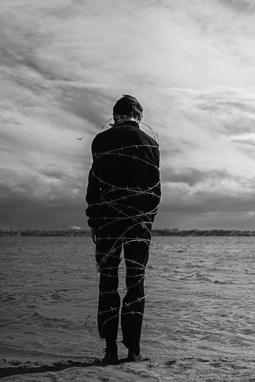 Man wrapped in barbed wire looks at a stormy sea under a cloudy sky in this high-contrast monochrome scene.