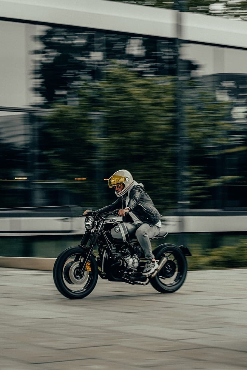Motorcyclist wearing a gold visor helmet and leather jacket speeds past blurred trees and buildings on a city street.