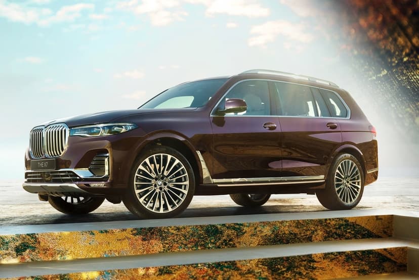 Burgundy BMW X7 SUV parked on a platform with chrome accents under a bright, hazy sky with lens flare.