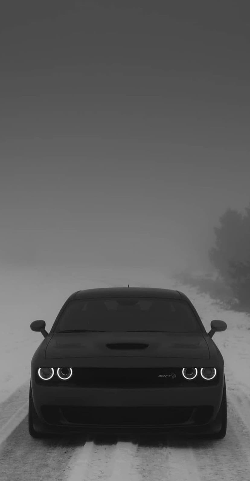 Black Dodge Challenger with glowing halo headlights parked on a snowy, fog-covered road during a cold winter day.