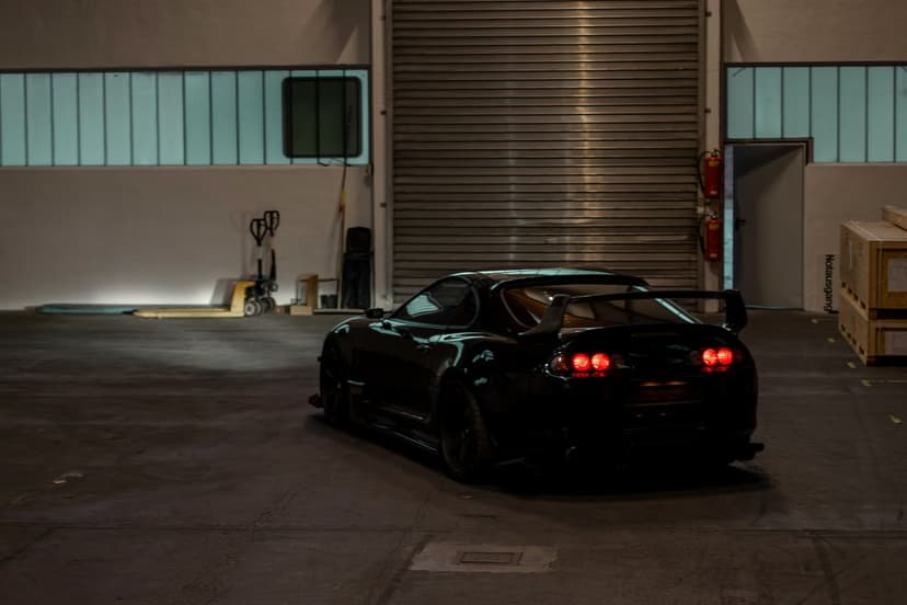 Toyota Supra rear view with red taillights in a dimly lit industrial warehouse with wooden crates.