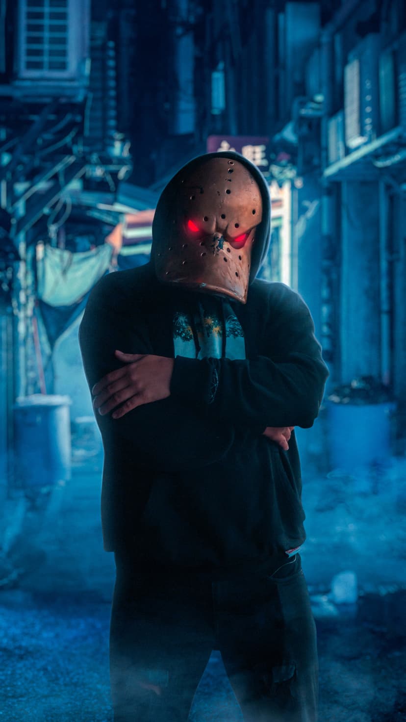 Masked figure with glowing red eyes and a menacing grin stands in a dark blue urban alleyway at night.