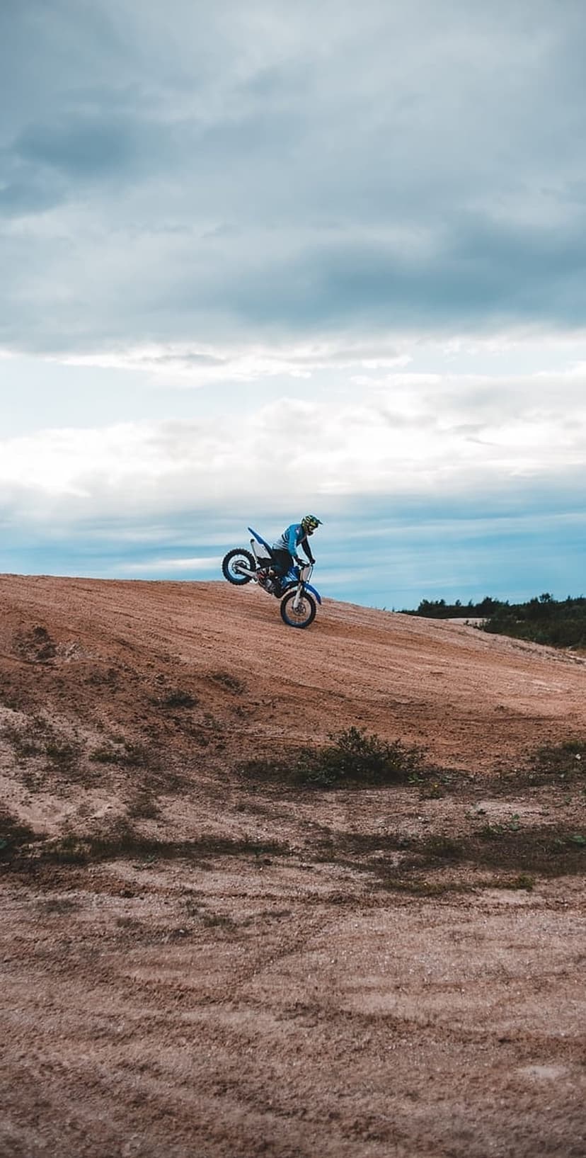 Motocross rider in blue gear performs a wheelie on a dusty track under a cloudy sky during an off-road race.